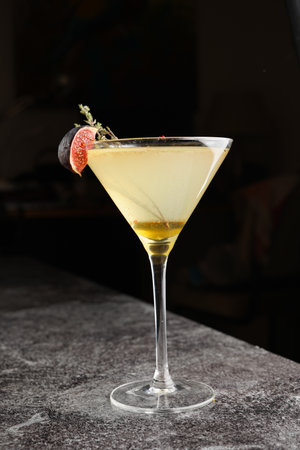 One martini glass with yellow alcoholic beverage liquor with thyme branch, honey and figs on gray table, dark bar backgroundの写真素材