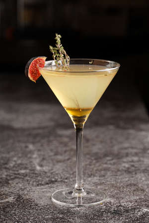 One martini glass with yellow alcoholic beverage liquor with thyme branch, honey and figs on gray table, dark bar backgroundの写真素材