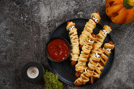 Cute Halloween party snack: wiener sausage with sugar eyes wrapped in dough stripes resembling mummies on a black plate with tomato ketchupの写真素材