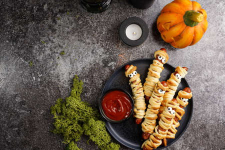 Cute Halloween party snack: wiener sausage with sugar eyes wrapped in dough stripes resembling mummies on a black plate with tomato ketchupの写真素材
