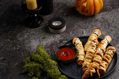 Cute Halloween party snack: wiener sausage with sugar eyes wrapped in dough stripes resembling mummies on a black plate with tomato ketchupの写真素材
