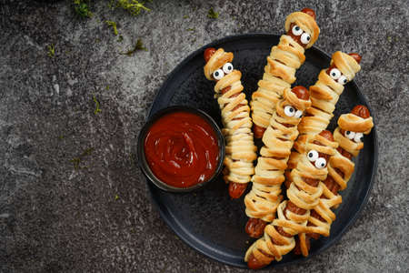 Cute Halloween party snack: wiener sausage with sugar eyes wrapped in dough stripes resembling mummies on a black plate with tomato ketchupの写真素材