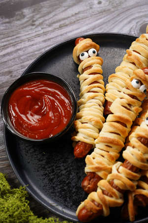 Cute Halloween party snack: wiener sausage with sugar eyes wrapped in dough stripes resembling mummies on a black plate with tomato ketchupの写真素材