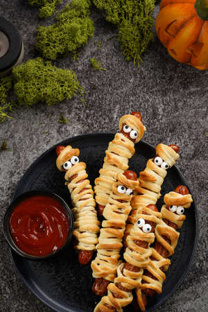 Cute Halloween party snack: wiener sausage with sugar eyes wrapped in dough stripes resembling mummies on a black plate with tomato ketchupの写真素材