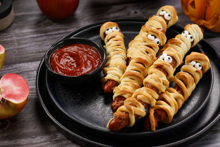 Cute Halloween party snack: wiener sausage with sugar eyes wrapped in dough stripes resembling mummies on a black plate with tomato ketchupの写真素材