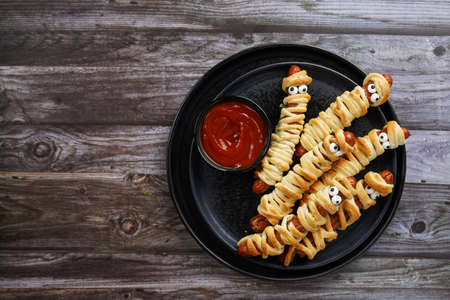 Cute Halloween party snack: wiener sausage with sugar eyes wrapped in dough stripes resembling mummies on a black plate with tomato ketchupの写真素材