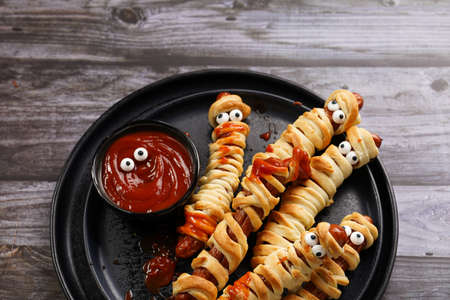 Cute Halloween party snack: wiener sausage with sugar eyes wrapped in dough stripes resembling mummies on a black plate with tomato ketchupの写真素材