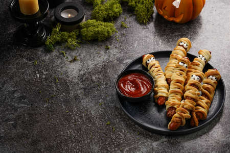 Cute Halloween party snack: wiener sausage with sugar eyes wrapped in dough stripes resembling mummies on a black plate with tomato ketchupの写真素材