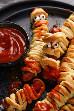 Cute Halloween party snack: wiener sausage with sugar eyes wrapped in dough stripes resembling mummies on a black plate with tomato ketchupの写真素材