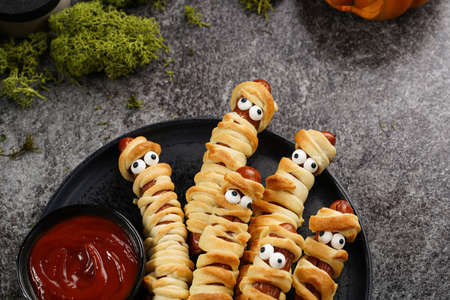 Cute Halloween party snack: wiener sausage with sugar eyes wrapped in dough stripes resembling mummies on a black plate with tomato ketchupの写真素材