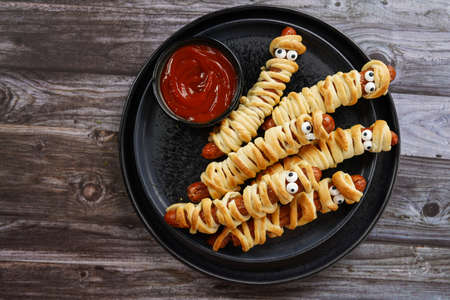 Cute Halloween party snack: wiener sausage with sugar eyes wrapped in dough stripes resembling mummies on a black plate with tomato ketchupの写真素材