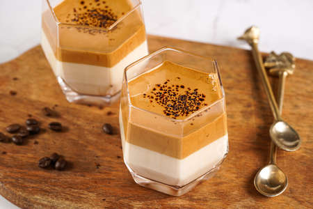 Two glasses with trendy korean beverage dalgona coffee latte - whipped instant coffee, sugar and hot water and milk layers on a wooden board, white surfaceの写真素材