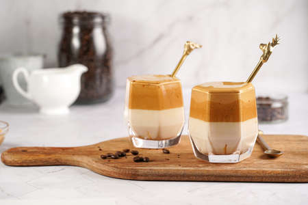 Two glasses with trendy korean beverage dalgona coffee latte - whipped instant coffee, sugar and hot water and milk layers on a wooden board, white surfaceの写真素材