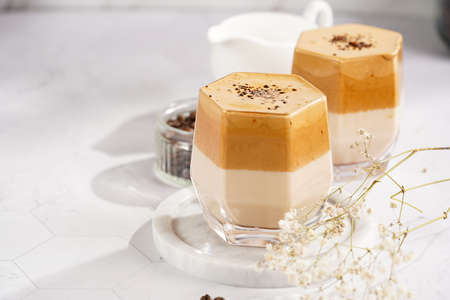 Two glasses with trendy korean beverage dalgona coffee latte - whipped instant coffee, sugar and hot water and milk layers on a marble board, white surfaceの写真素材