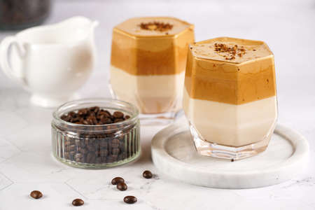 Two glasses with trendy korean beverage dalgona coffee latte - whipped instant coffee, sugar and hot water and milk layers on a wooden board, white surfaceの写真素材