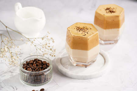 Two glasses with trendy korean beverage dalgona coffee latte - whipped instant coffee, sugar and hot water and milk layers on a marble board, white surfaceの写真素材