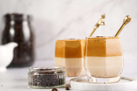 Two glasses with trendy korean beverage dalgona coffee latte - whipped instant coffee, sugar and hot water and milk layers on a wooden board, white surfaceの写真素材