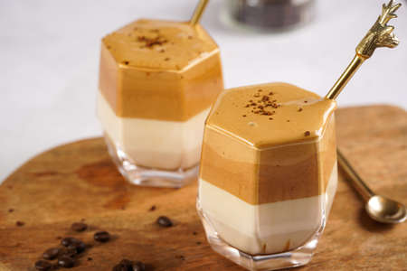 Two glasses with trendy korean beverage dalgona coffee latte - whipped instant coffee, sugar and hot water and milk layers on a wooden board, white surfaceの写真素材