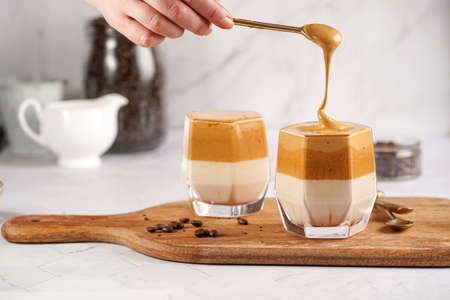 Female hand stirring trendy korean beverage dalgona coffee latte - whipped instant coffee, sugar and hot water and milk layers on a marble board, white surfaceの写真素材