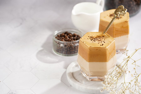 Two glasses with trendy korean beverage dalgona coffee latte - whipped instant coffee, sugar and hot water and milk layers on a marble board, white surfaceの写真素材