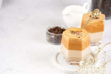 Two glasses with trendy korean beverage dalgona coffee latte - whipped instant coffee, sugar and hot water and milk layers on a marble board, white surfaceの写真素材