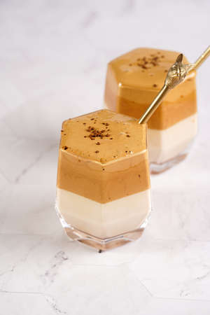 Two glasses with trendy korean beverage dalgona coffee latte - whipped instant coffee, sugar and hot water and milk layers on a wooden board, white surfaceの写真素材