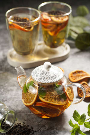 A transparent tea pot filled with black tea, oranges and mint on a gray surfaceの写真素材