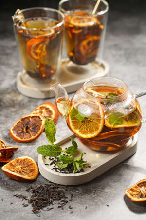 A transparent tea pot filled with black tea, oranges and mint on a gray surfaceの写真素材