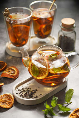 A transparent tea pot filled with black tea, oranges and mint on a gray surfaceの写真素材