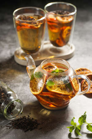A transparent tea pot filled with black tea, oranges and mint on a gray surfaceの写真素材