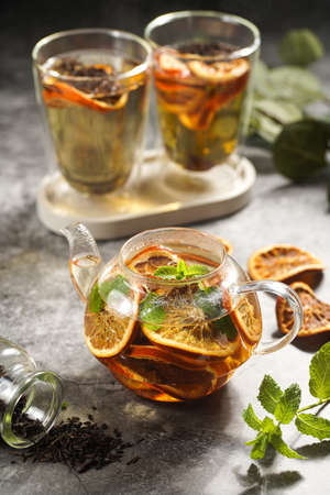 A transparent tea pot filled with black tea, oranges and mint on a gray surfaceの写真素材