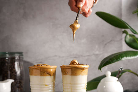 Male hand stirring trendy korean beverage dalgona coffee latte - whipped instant coffee, sugar and hot water and milk layers on a marble board, gray surfaceの写真素材
