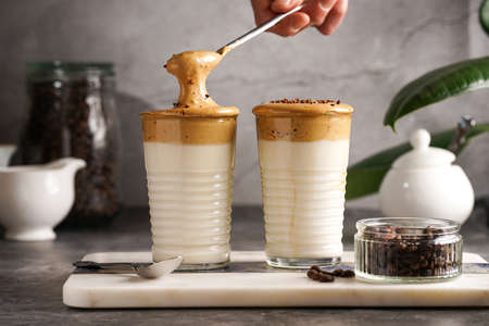 Male hand stirring trendy korean beverage dalgona coffee latte - whipped instant coffee, sugar and hot water and milk layers on a marble board, gray surfaceの写真素材