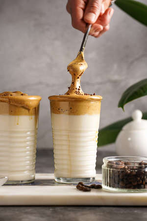 Male hand stirring trendy korean beverage dalgona coffee latte - whipped instant coffee, sugar and hot water and milk layers on a marble board, gray surfaceの写真素材