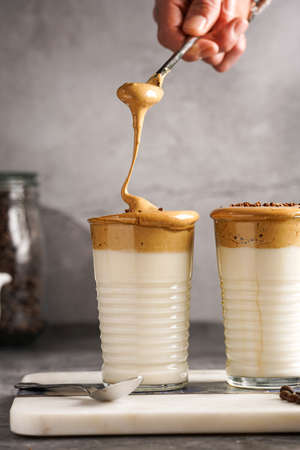 Male hand stirring trendy korean beverage dalgona coffee latte - whipped instant coffee, sugar and hot water and milk layers on a marble board, gray surfaceの写真素材