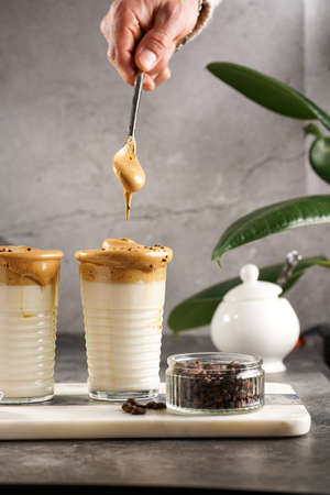 Male hand stirring trendy korean beverage dalgona coffee latte - whipped instant coffee, sugar and hot water and milk layers on a marble board, gray surfaceの写真素材