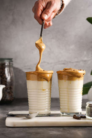 Male hand stirring trendy korean beverage dalgona coffee latte - whipped instant coffee, sugar and hot water and milk layers on a marble board, gray surfaceの写真素材