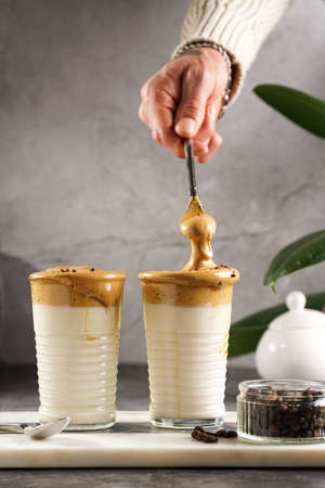 Male hand stirring trendy korean beverage dalgona coffee latte - whipped instant coffee, sugar and hot water and milk layers on a marble board, gray surfaceの写真素材