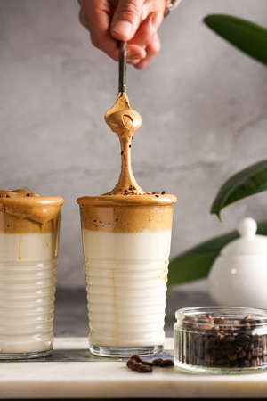 Male hand stirring trendy korean beverage dalgona coffee latte - whipped instant coffee, sugar and hot water and milk layers on a marble board, gray surfaceの写真素材