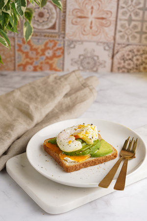 Savory breakfast - A toast bread with cream cheese, avocado and poached egg on a white plate on marble trayの写真素材