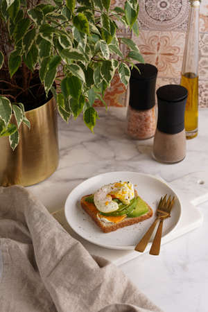 Savory breakfast - A toast bread with cream cheese, avocado and poached egg on a white plate on marble trayの写真素材