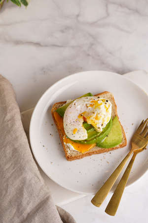 Savory breakfast - A toast bread with cream cheese, avocado and poached egg on a white plate on marble trayの写真素材