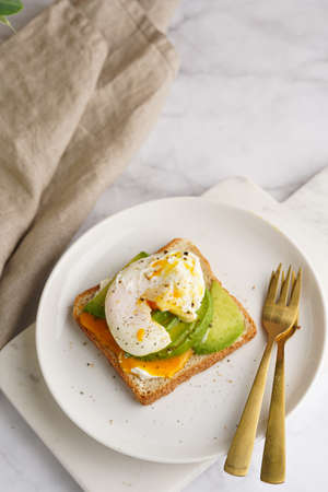 Savory breakfast - A toast bread with cream cheese, avocado and poached egg on a white plate on marble trayの写真素材
