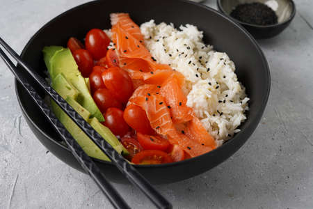 Poke bowl - black bowl with white rice, smoked salmon, cherry tomatoes and avocado, sesame seeds, soy sauce and chopsticks on a gray concrete surfaceの写真素材