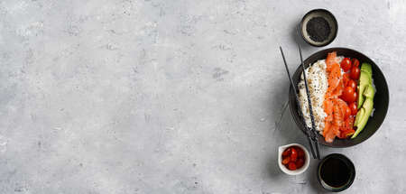 Poke bowl - black bowl with white rice, smoked salmon, cherry tomatoes and avocado, sesame seeds, soy sauce and chopsticks on a gray concrete surface. Top view, negative space, banner sizeの写真素材