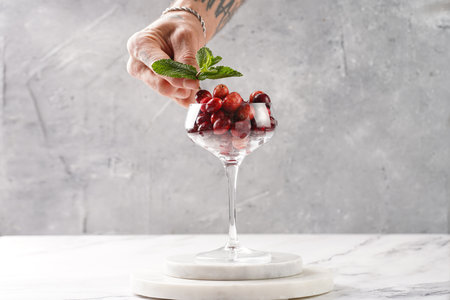 Male hand garnishing wide champagne glass filled with raw cranberries with fresh mint on a round marble trayの写真素材