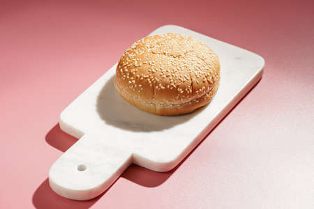 One white wheat burger bun with sesame seeds on marble board on pink seamless surface, hard lightの写真素材
