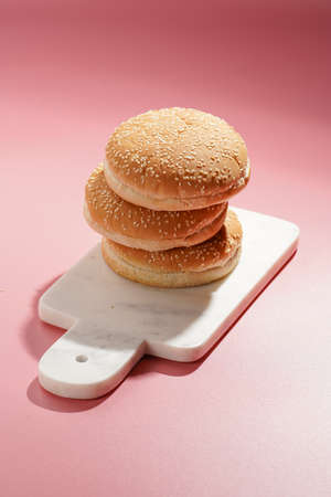 Three white wheat burger buns with sesame seeds on marble board on pink seamless surface, hard lightの写真素材