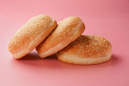 Three white wheat burger buns with sesame seeds on pink seamless surface