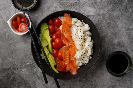 Poke bowl - black bowl with white rice, smoked salmon, cherry tomatoes and avocado, sesame seeds, soy sauce and chopsticks on a gray concrete surface, top viewの写真素材